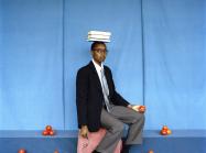 artist sits with books on head, surrounded by piles of tomatoes