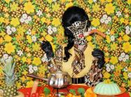 Miss meatface in neutral oranges and a leopard-print latex mask, sits in front of table loaded with pineapples, jello molds, and sausage plates. In front of busy floral background. 