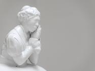 White material cast to show black female figure, dressed in clothes that seem to be from the 1800s, her hands are clasped together as if in prayer but held just to the side of her face to support her chin. 