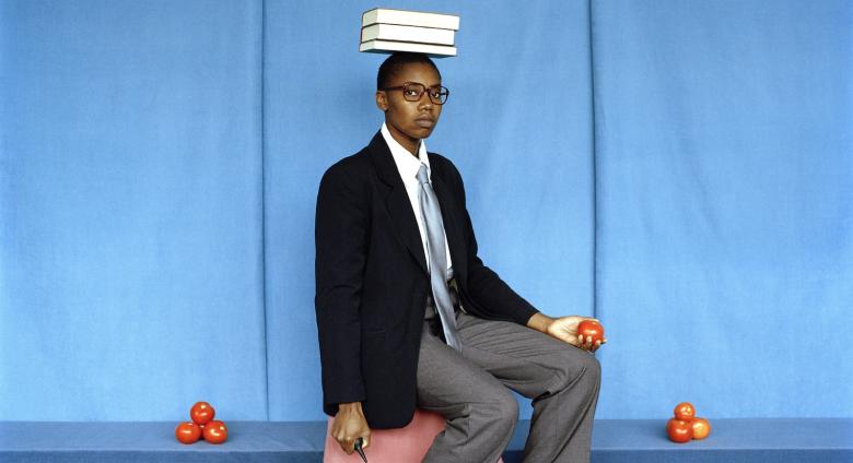 artist sits with books on head, surrounded by piles of tomatoes