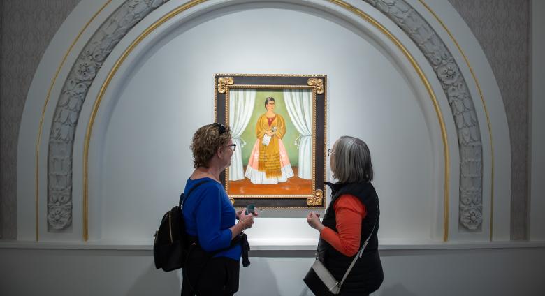 Two museum visitors are viewing a painting. The painting features a woman with a medium skin-tone wearing a dress and scarf and holding a piece of paper. It is framed by an arc with golden and floral ornamentation.