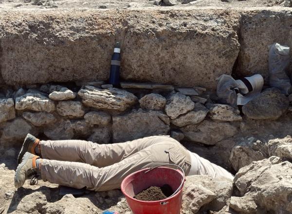 Excavation of certain physical features often requires archaeologists to get creative with digging methodologies. Wells, cisterns, and other subterranean features can be particularly challenging. Archeologist shown with head and torso in a hole as they work on site. 