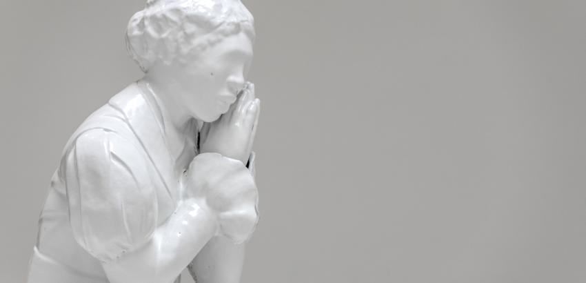 White material cast to show black female figure, dressed in clothes that seem to be from the 1800s, her hands are clasped together as if in prayer but held just to the side of her face to support her chin. 